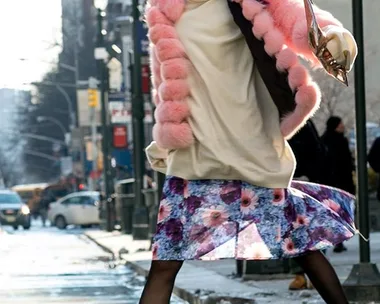 A woman in a pink fur coat and floral dress confidently strides down a snowy city street.