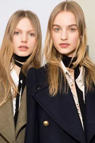 Two women with long, straight, blond hair wearing black neck scarves and fashionable coats, posing together.