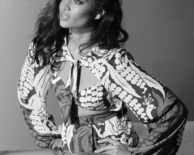 Fashion model poses in a patterned dress, seated, looking to the side. Black and white portrait.