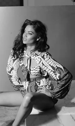 Fashion model poses in a patterned dress, seated, looking to the side. Black and white portrait.