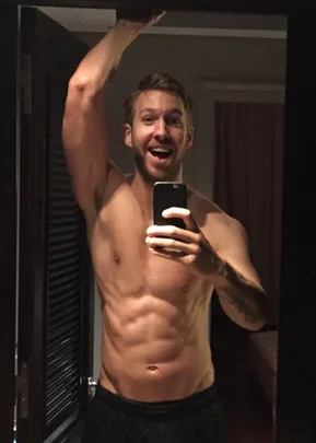 Man taking a mirror selfie, shirtless, displaying a toned physique, with a joyful expression.