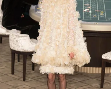 Model in a feathered ivory dress walks at Chanel Haute Couture AW15/16 runway show, set around a casino table.