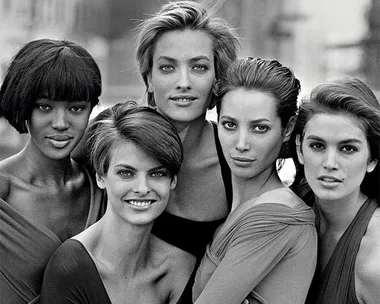 Black and white photo of five supermodels posing together, smiling, in a city setting.