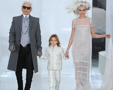 Three models walk down a runway: an elderly man in gray, a child in white, and a woman in a sheer dress with a feathered headpiece.