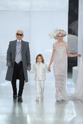 Three models walk down a runway: an elderly man in gray, a child in white, and a woman in a sheer dress with a feathered headpiece.