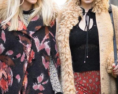 Two women wearing 70s inspired fashion with sunglasses and patterned coats walking outdoors.