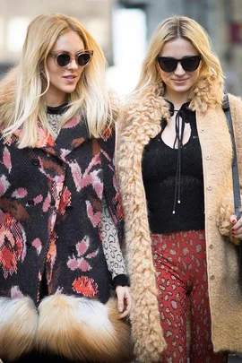 Two women wearing 70s inspired fashion with sunglasses and patterned coats walking outdoors.