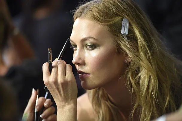 Woman applying makeup with a brush, hair clipped back, focused expression, in a backstage setting.