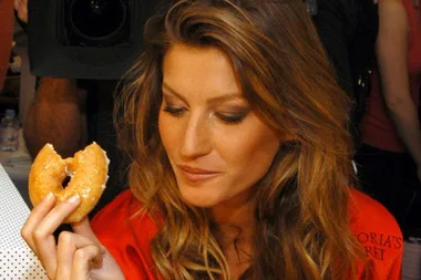 A woman with long brown hair holds a glazed doughnut, wearing a red Victoria's Secret robe, looking at the doughnut.