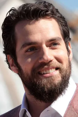 A smiling man with dark hair and a full beard, wearing a light-colored shirt and brown blazer.