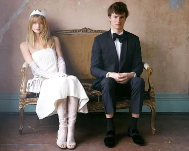 A woman in a white dress and gloves with a crown and a man in a black suit and bow tie sitting on a vintage sofa.