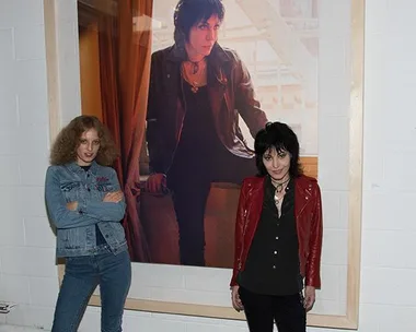 Two women stand in front of a large framed photo; one in a denim outfit, the other in a red jacket.