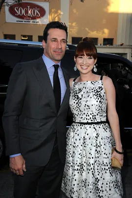 Jon Hamm in a suit and Ellie Kemper in a patterned dress, smiling and standing together outside in front of a Socko's Subs sign.