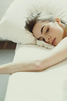 A woman with light hair is lying on her side on a bed, resting her head on a white pillow, with her arm outstretched.