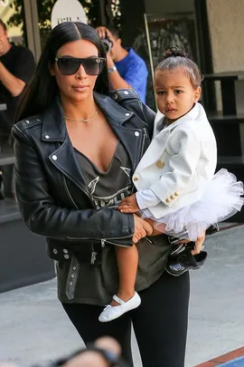 Kim Kardashian carrying daughter North West, both wearing stylish clothes and sunglasses, outside in daylight.