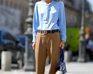 Woman in a light blue blouse, brown trousers, sunglasses, holding a blue handbag with a fur accessory, standing on a city street.