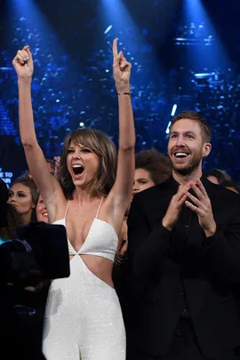 Two people celebrating at an event, with one in a white outfit raising their arms and the other in black clapping.