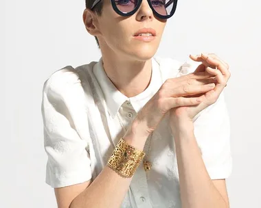 "Person with short dark hair wearing large black sunglasses, a white shirt, and a gold bracelet, hands clasped together on a table."