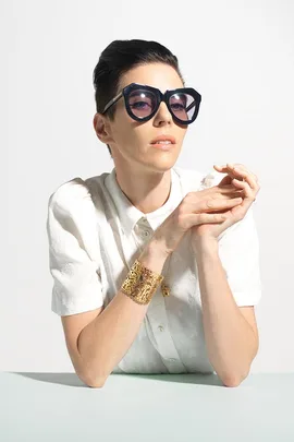 "Person with short dark hair wearing large black sunglasses, a white shirt, and a gold bracelet, hands clasped together on a table."