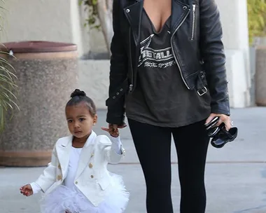 Woman in leather jacket and sunglasses holding hand of a young girl in a white dress and jacket walking outdoors.