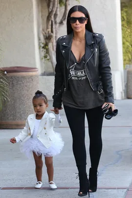 Woman in leather jacket and sunglasses holding hand of a young girl in a white dress and jacket walking outdoors.