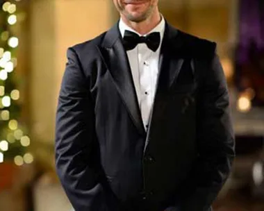 Man in black tuxedo and bow tie, standing outside with a tree decorated with lights in the background.