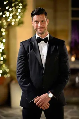 Man in black tuxedo and bow tie, standing outside with a tree decorated with lights in the background.