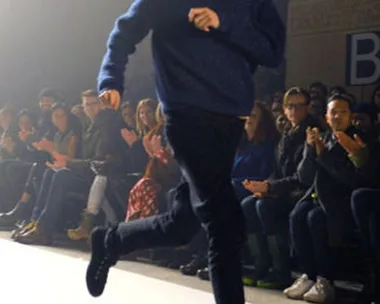 Designer running on the runway during a fashion show, with audience clapping in the background.