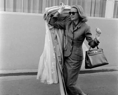 A woman in a suit and sunglasses carries a dress over her shoulder, smiling as she walks outdoors.