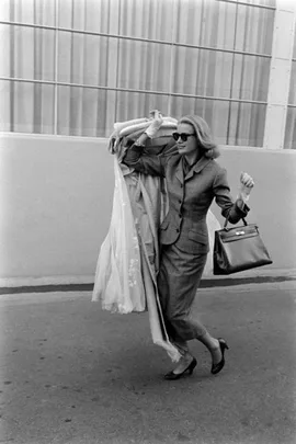 A woman in a suit and sunglasses carries a dress over her shoulder, smiling as she walks outdoors.