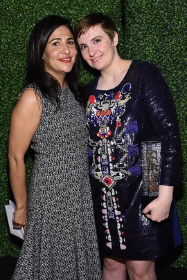 Two women posing together; one in a patterned sleeveless dress and the other in a sequined dress, both smiling.