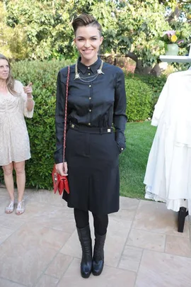 Ruby Rose in an all-black outfit with a red purse, standing outdoors at an event, smiling at the camera.