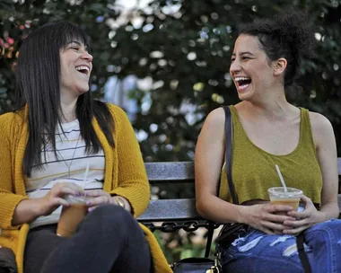 Two women laughing and sitting on a bench, holding iced drinks on a sunny day.