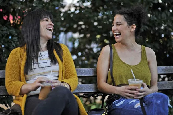 Two women laughing and sitting on a bench, holding iced drinks on a sunny day.