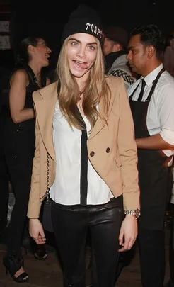 A woman in a beige jacket and beanie making a funny face with her tongue out, surrounded by people at an event.