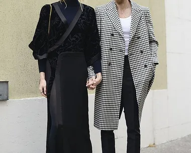 A couple holding hands, dressed in stylish outfits; the woman in a black dress with pink sneakers, the man in a black-and-white coat.