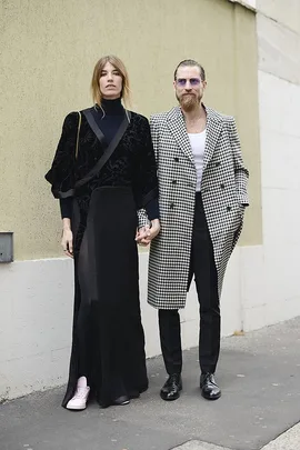 A couple holding hands, dressed in stylish outfits; the woman in a black dress with pink sneakers, the man in a black-and-white coat.