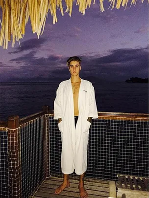 Young man in a white robe standing on a deck beside the sea during sunset, under a thatched roof.