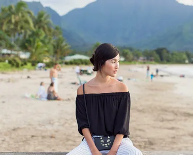 Woman sitting on a beach wall, wearing an off-shoulder top and striped pants, scenic mountains in the background.