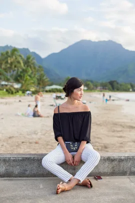 Woman sitting on a beach wall, wearing an off-shoulder top and striped pants, scenic mountains in the background.