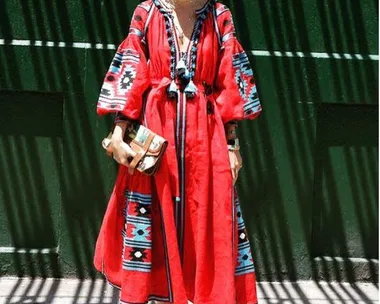 Person wearing a red bohemian dress with blue and black patterns, holding a clutch, standing against a green wall.