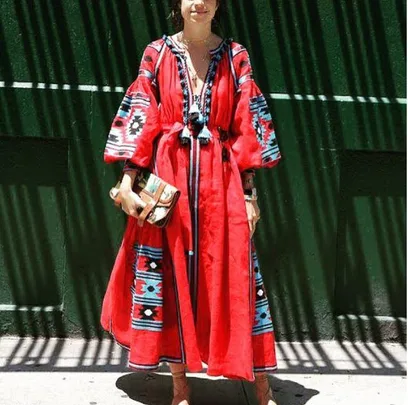 Person wearing a red bohemian dress with blue and black patterns, holding a clutch, standing against a green wall.