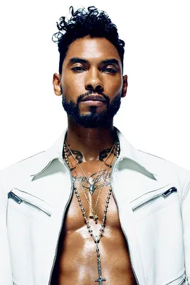 Miguel, shirtless, wears a white jacket and layered necklaces in a close-up, high-contrast portrait for an Elle interview.