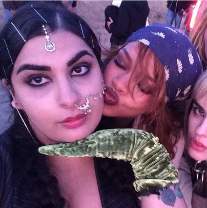 Three women pose for a photo, one with face jewelry and another kissing her cheek playfully, with a green glove visible.