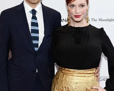 A man in a navy suit and glasses poses with a woman in a black top and gold skirt at a formal event backdrop.