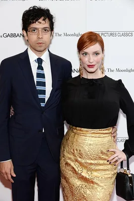 A man in a navy suit and glasses poses with a woman in a black top and gold skirt at a formal event backdrop.