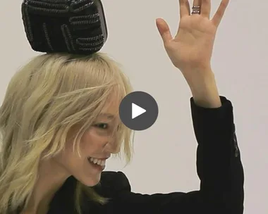 Model with blonde hair, smiling and waving, while balancing a black purse on her head.