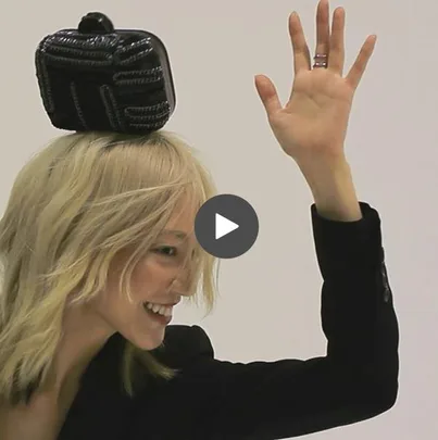 Model with blonde hair, smiling and waving, while balancing a black purse on her head.