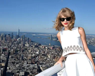 Taylor Swift in white outfit and sunglasses, posing on a rooftop with a city skyline and river in the background.