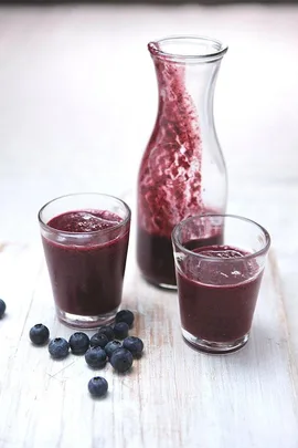 Three glasses of blueberry smoothie with a glass carafe and fresh blueberries on a wooden surface.